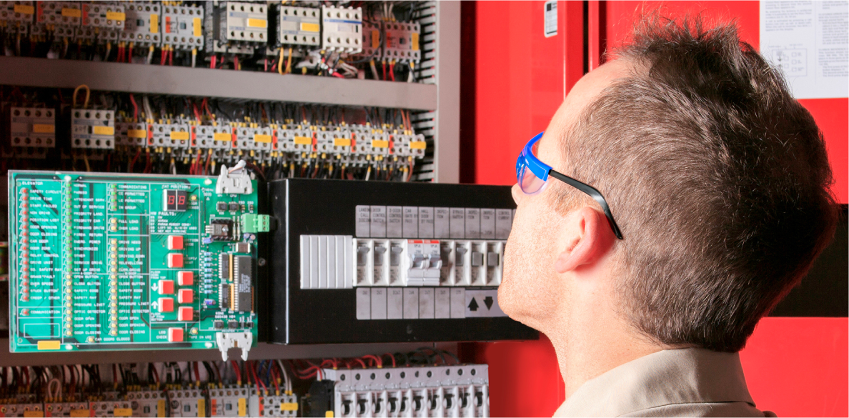 Technician wearing safety glasses inspects electrical control panel with circuit breakers and wiring in an industrial setting.