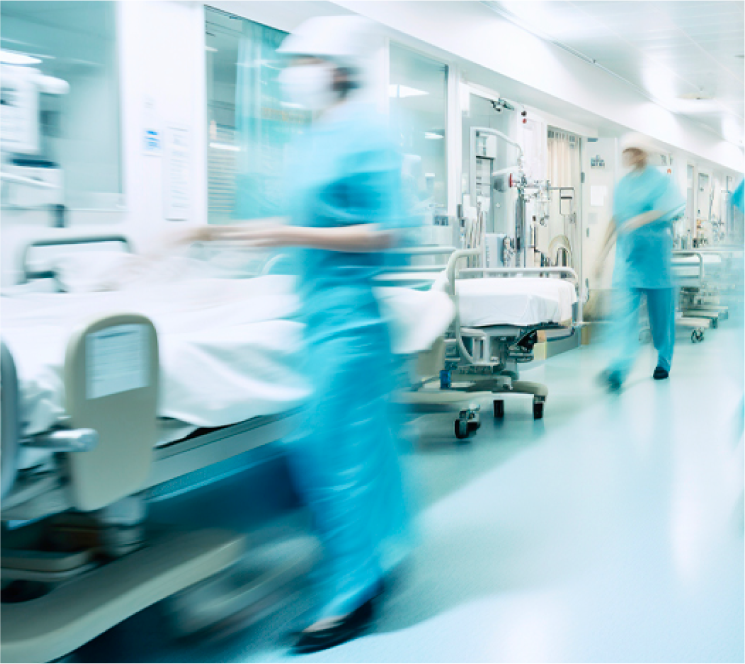 Blurred medical staff in blue uniforms moving through a bright hospital corridor with patient beds and medical equipment.