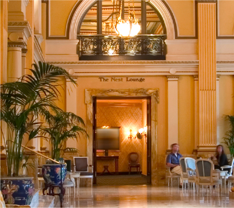 Elegant hotel lobby with classical architecture, potted plants, and guests seated in a lounge area.