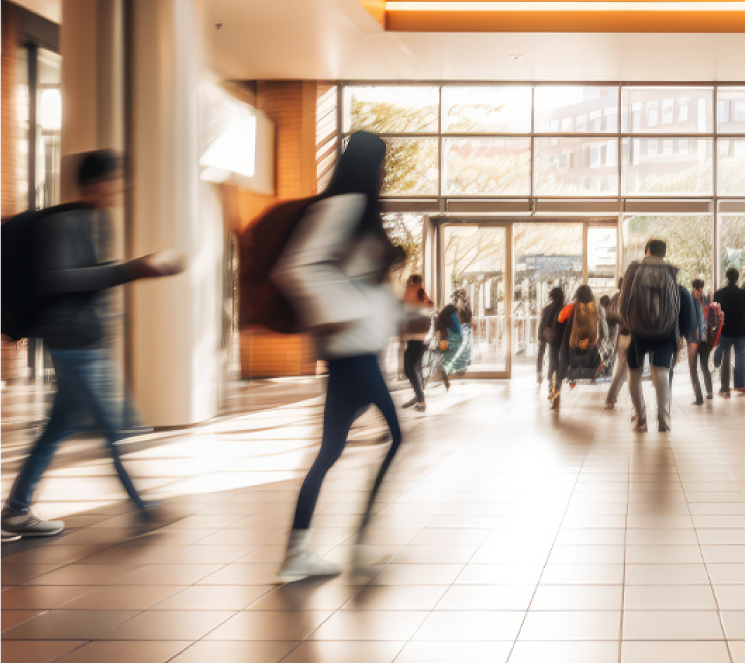 Multiple people moving through a bright, modern building lobby with large windows and tiled flooring.