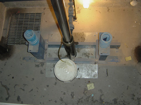 Overhead view of elevator machinery room floor with metal components, pipes, and blue cylindrical equipment on concrete.