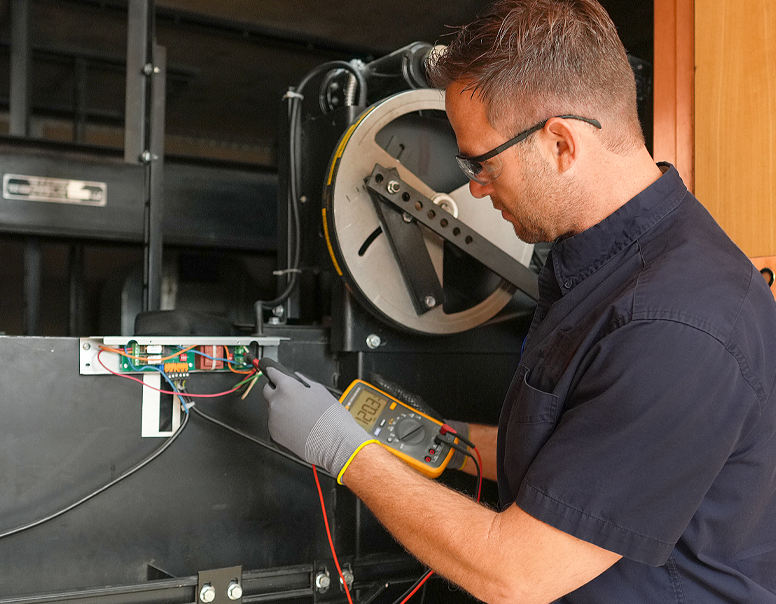 Technician wearing safety glasses uses a multimeter to test electrical components on an elevator control panel.