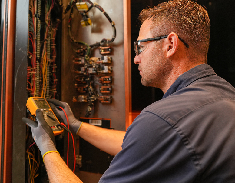 Technician wearing safety glasses using a multimeter to test electrical components inside an elevator control panel.