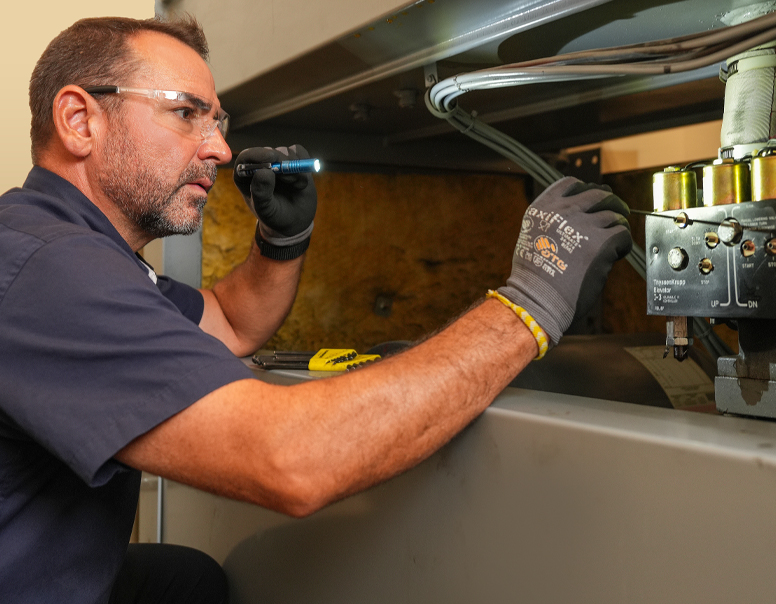 Technician in safety glasses inspecting elevator machinery and wiring with a flashlight.