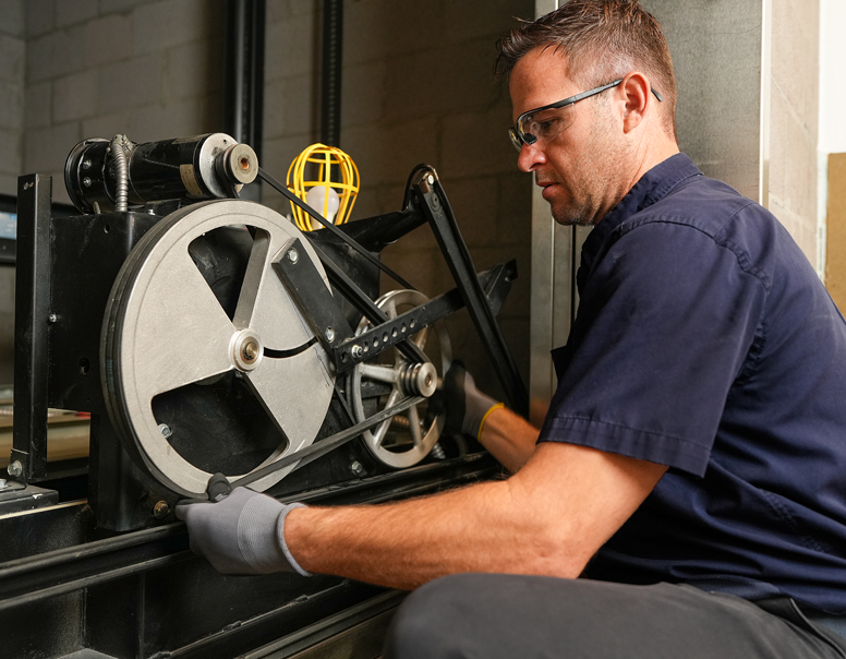 Man wearing safety glasses and navy shirt adjusts machinery with a large wheel and yellow safety cage.