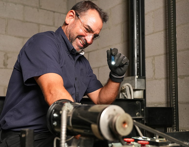 Technician in dark blue shirt wearing black gloves inspecting elevator equipment with a flashlight.