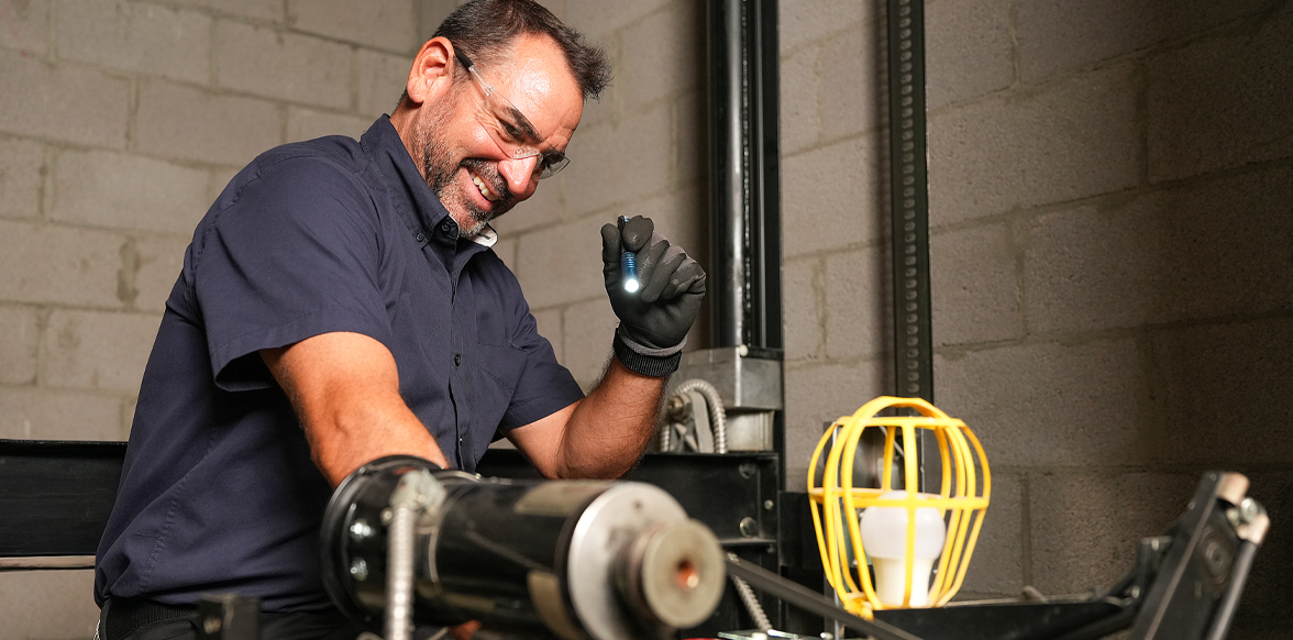 Technician in navy shirt wearing black gloves inspects elevator mechanical components with a headlamp.