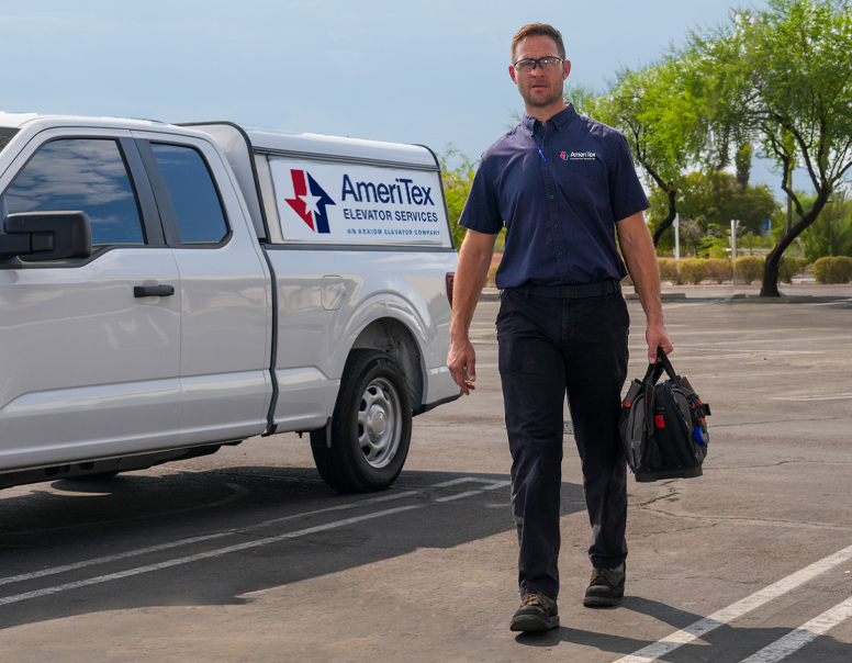 Service technician in company uniform walking with tools next to AmerTex Elevator Services van in parking lot.